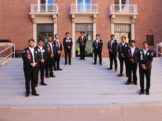 Chapter photo of Omega Delta Phi. They're standing on steps in a triangular formation wearing black pants and line jackets.